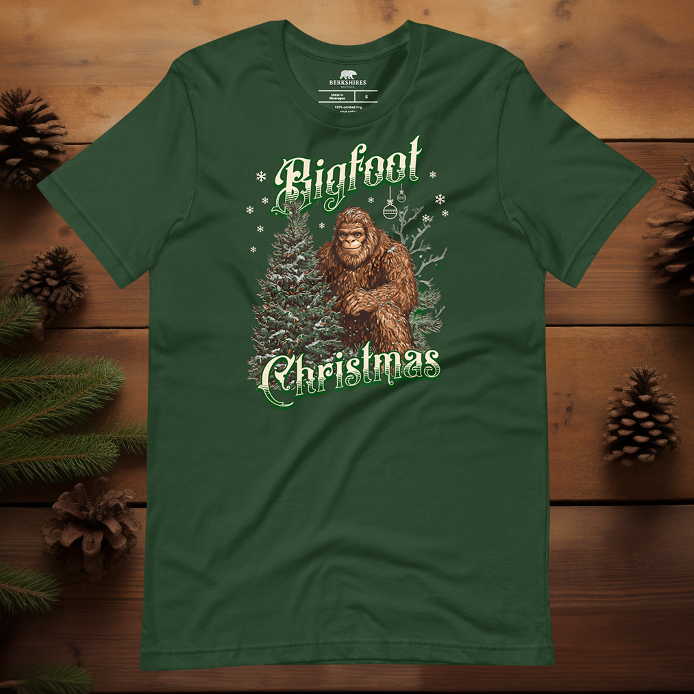 Forest green Sasquatch Christmas t-shirt with Bigfoot beside a holiday tree under falling snow, featuring retro green “Bigfoot Christmas” lettering for nature lovers and cryptid fans.
