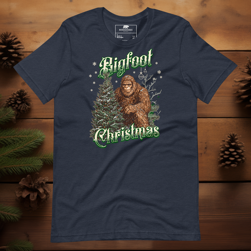 Heather navy Bigfoot Christmas tee with a detailed illustration of Bigfoot in front of pine trees, surrounded by snowflakes and ornaments, with green gothic-style holiday text.
