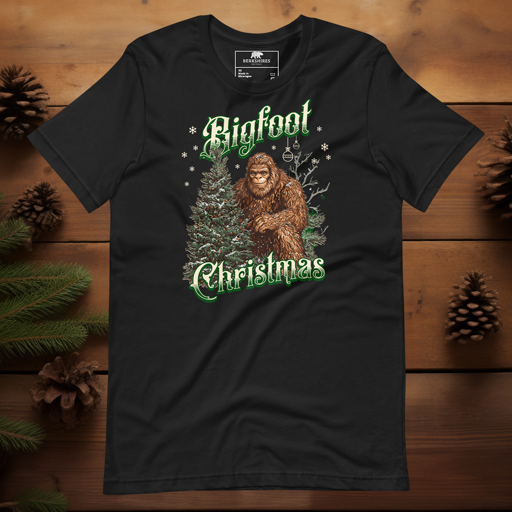 Bigfoot Christmas t-shirt featuring a friendly Bigfoot standing beside a decorated evergreen tree with snowflakes falling, text reads “Bigfoot Christmas” in green and white vintage lettering on a black tee.