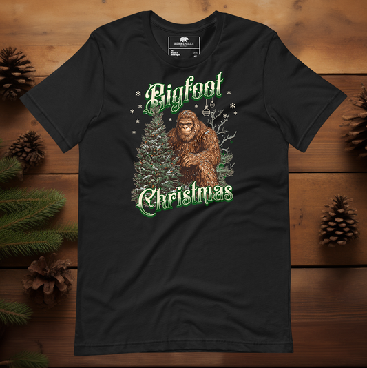 Bigfoot Christmas t-shirt featuring a friendly Bigfoot standing beside a decorated evergreen tree with snowflakes falling, text reads “Bigfoot Christmas” in green and white vintage lettering on a black tee.